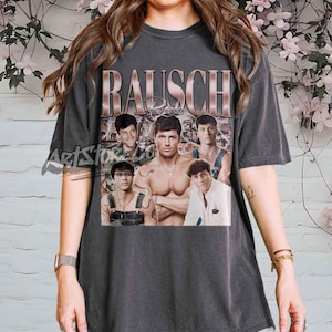 May include: Dark gray t-shirt featuring a vintage-style graphic with the name "RAUSCH" in a copper-colored font. The graphic includes multiple images of a man in various poses. The shirt is a casual, oversized style.