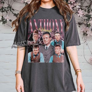 May include: Dark gray t-shirt featuring a vintage-style graphic of Anthony Head. The design includes the name "ANTHONY Head" in a pink and gold font, with multiple images of the actor. The shirt is a classic crew neck.