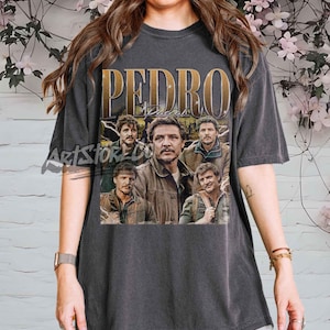 May include: Dark grey t-shirt with the name "PEDRO" in gold lettering and a collage of images of a man. The images are in various poses. The t-shirt is a casual, unisex style.