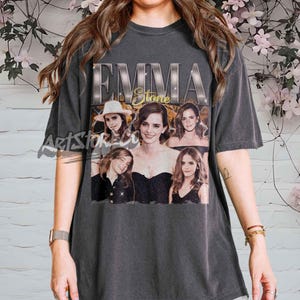 Puede incluir: Camiseta gris oscuro con un gráfico vintage de Emma Stone, con el texto "EMMA Stone" y varias imágenes de la actriz. La camiseta tiene mangas cortas y cuello redondo.
