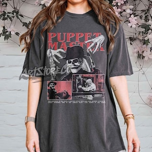May include: Dark gray t-shirt featuring a graphic from the movie "Puppet Master." The design includes the text "Puppet Master" in red, a skull-faced character in a hat, and other imagery from the film. The shirt also has additional text.