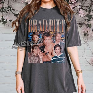 Può includere: T-shirt grigio scuro con una grafica di Brad Pitt e la scritta "BRAD PITT" in arancione e oro. La grafica include diverse immagini dell'attore. La maglietta ha maniche corte e girocollo.