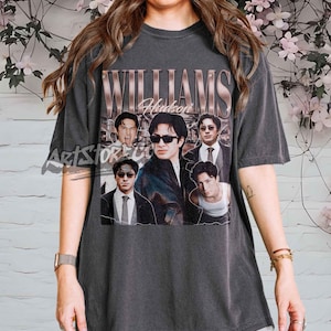 May include: Dark gray t-shirt featuring a vintage-style graphic with the name "WILLIAMS Hudson" and multiple images of a man. The design has a faded, distressed look. The shirt is short-sleeved and appears to be made of a soft material.