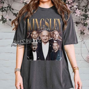 Puede incluir: Camiseta gris oscuro con un gráfico dorado con el nombre "KINGSLEY" y las imágenes de un hombre. La camiseta tiene un estilo vintage y una apariencia ligeramente descolorida, lo que le da un aspecto desgastado.