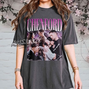 Può includere: T-shirt grigio scuro con una grafica con la scritta "CHENFORD" in un font sfumato rosa e bianco. La grafica include più immagini di persone. La maglietta è in stile vintage.