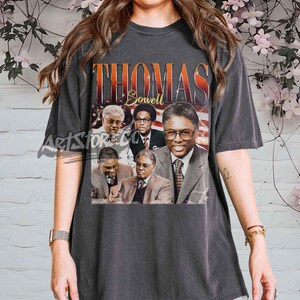 May include: Dark grey t-shirt with a graphic of Thomas Sowell and the text "THOMAS SOWELL" in orange and red. The graphic includes multiple images of Thomas Sowell in suits. The shirt has a vintage style.