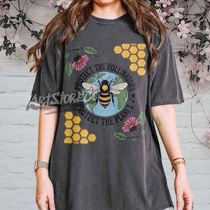 May include: Dark gray t-shirt with a graphic featuring a bee over a globe, surrounded by text that reads "Protect the Pollinators, Protect the Planet." The design includes honeycomb and floral elements.
