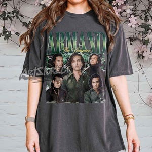 May include: Dark gray t-shirt featuring a graphic of the word "ARMAND" in green, with the text "The Vampire" below. The shirt displays a collage of images of a person. The shirt is a vintage style.