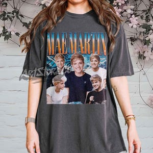Op de afbeelding: Donkergrijs t-shirt met een retro graphic van Mae Martin en de tekst "MAE MARTIN". Het ontwerp bevat meerdere afbeeldingen van Mae Martin. Het shirt heeft een vintage, versleten uitstraling.
