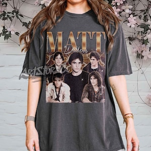 Puede incluir: Camiseta gris oscuro con un gráfico de estilo vintage. El gráfico presenta la palabra "MATT" en letras doradas e imágenes de varias personas. La camiseta tiene cuello redondo y mangas cortas.