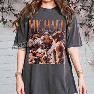Könnte beinhalten: Dunkelgraues T-Shirt mit einer Grafik von Michael B. Jordan und dem Text "MICHAEL B. Jordan" in Orange und Weiß. Die Grafik enthält Bilder des Schauspielers. Das T-Shirt hat kurze Ärmel.