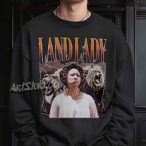 May include: Black sweatshirt featuring a graphic of a woman with lions and the text "LAND LADY" in orange and white. The image has a vintage, distressed aesthetic with lightning bolts in the background.