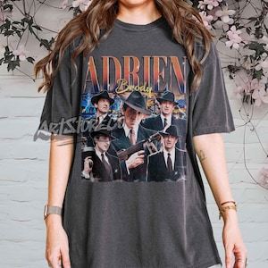 May include: Dark gray t-shirt featuring a vintage-style graphic of actor Adrien Brody in a fedora and suit. The shirt has the name "Adrien Brody" printed in a retro font. The shirt is a short-sleeved, crew neck style.