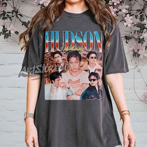 May include: Dark gray t-shirt featuring a vintage-style graphic with the name "HUDSON" in blue and red lettering. The graphic includes multiple images of a person and the word "Real".