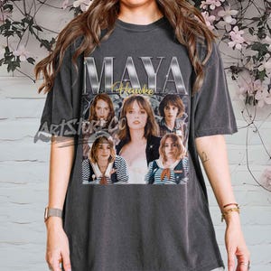 Könnte beinhalten: Dunkelgraues T-Shirt mit einer Collage von Maya Hawke-Bildern, dem Namen "MAYA" in großen silbernen Buchstaben und "Hawke" in goldener Schrift. Das T-Shirt hat einen Vintage-Look.