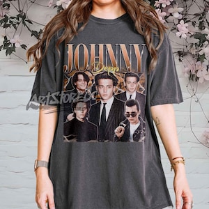 May include: Dark gray t-shirt featuring a vintage-style graphic of Johnny Depp. The design includes the name "JOHNNY Depp" in gold lettering above a collage of Depp's images. The shirt has short sleeves and a crew neck.
