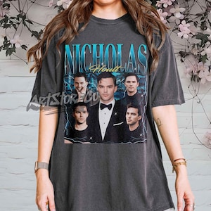 May include: Dark gray t-shirt featuring a vintage-style graphic of Nicholas Hoult with the text "Nicholas Hoult" in a blue and white design. The shirt has a crew neck and short sleeves, and the graphic includes multiple images of the actor.