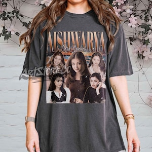 Puede incluir: Camiseta gris oscuro con un gráfico vintage de Aishwarya Rai Bachchan. El diseño incluye múltiples imágenes de la actriz con el nombre "Aishwarya Rai Bachchan" en una fuente dorada retro. La camiseta tiene mangas cortas y cuello redondo.