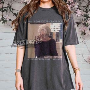 Könnte beinhalten: Dunkelgraues T-Shirt mit einem rechteckigen Bild einer Frau mit Brille und dem Text "BY ALL MEANS MOVE AT A GLACIAL PACE, YOU KNOW HOW THAT THRILLS ME." Das T-Shirt hat kurze Ärmel und eine lockere Passform.