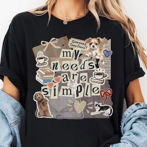 May include: Black t-shirt with a collage design featuring the text "my needs are simple." The design includes images of a dog, cat, books, coffee cups, and hearts. The shirt is worn with a denim jacket and shorts.