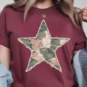 May include: A maroon t-shirt featuring a large star design. The star is made of patchwork fabric in shades of green, pink, and beige, with a distressed, frayed edge. The shirt has short sleeves and a crew neck.