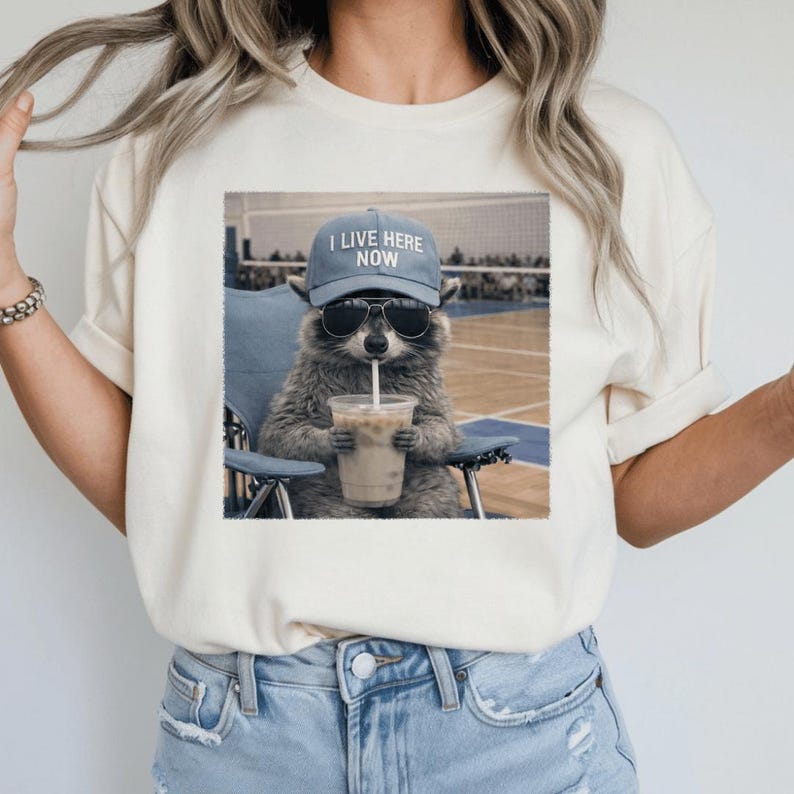 May include: Cream-colored t-shirt featuring a raccoon wearing sunglasses and a blue cap with the text "I LIVE HERE NOW." The raccoon is sitting in a blue chair and drinking from a clear cup with a straw.