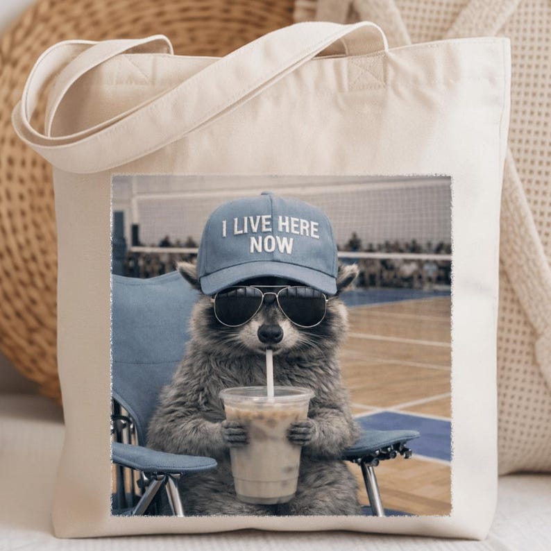 May include: Beige tote bag featuring a graphic of a raccoon wearing sunglasses and a blue cap that reads "I LIVE HERE NOW." The raccoon is drinking from a clear cup with a straw while sitting in a blue chair.