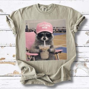 May include: A light olive green t-shirt featuring a graphic of a raccoon wearing sunglasses and a pink hat that says "I LIVE HERE NOW." The raccoon is sitting in a pink chair and drinking an iced coffee.