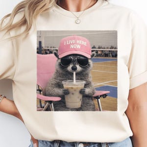 May include: Cream-colored t-shirt featuring a raccoon wearing sunglasses and a pink cap that reads "I LIVE HERE NOW." The raccoon is sitting in a pink chair and drinking from a cup with a straw. The image has a vintage, slightly faded look.