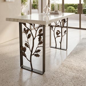 May include: A rectangular table with a white marble top and two dark metal legs. The legs feature a decorative design of tree branches with small oak leaves. The table is positioned in a bright room with large glass windows and doors.
