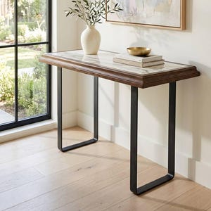 May include: A rectangular console table featuring a white marble top with grey veining, a dark brown wooden frame, and two black metal U-shaped legs. The table holds a cream-colored ceramic vase with green branches, two stacked books, and a gold bowl.