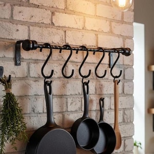 Puede incluir: Un estante de metal negro para ollas con seis ganchos curvos, montado en una pared de ladrillo. Tres sartenes negras y una cuchara de madera cuelgan de los ganchos. Una solución de almacenamiento de cocina.