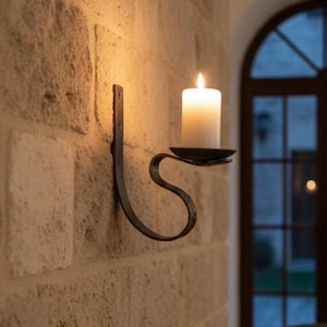 Könnte beinhalten: Ein schwarzer Wandkerzenhalter aus Metall mit einer brennenden weißen Kerze. Der Halter hat ein geschwungenes, dekoratives Design und ist an einer strukturierten Steinmauer befestigt. Der Kerzenhalter ist ein dekoratives Wohnaccessoire.