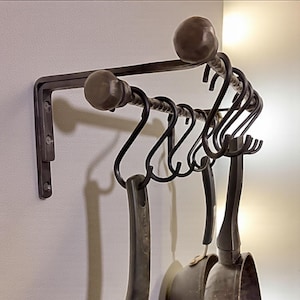 May include: A dark brown, wrought iron pot rack mounted on a wall. The rack features a horizontal bar with several S-shaped hooks for hanging cookware. Two decorative finials and a supporting bracket are also visible.