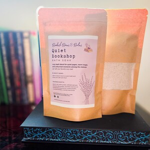 Peut inclure: Deux sachets en papier brun de bain moussant, l'un avec une étiquette « Quiet Bookshop BATH SOAK » et une illustration de lavande. L'autre sachet est partiellement transparent, révélant le produit. Les sachets sont posés sur une boîte noire décorative.