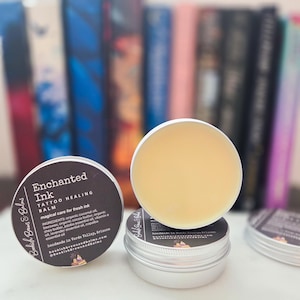 Puede incluir: Primer plano de tres pequeños botes plateados de bálsamo curativo para tatuajes. Uno de los botes está abierto, revelando un bálsamo amarillo claro. Las etiquetas dicen "Enchanted Ink Tattoo Healing Balm" con una lista de ingredientes. El fondo está borroso, mostrando libros coloridos.