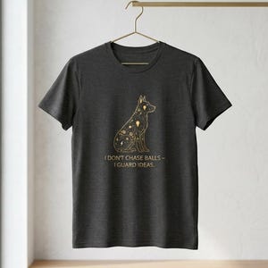 Może przedstawiać: Ciemnoszara koszulka z złotą ilustracją psa i napisem "I DON'T CHASE BALLS - I GUARD IDEAS." Projekt psa zawiera niebieskie i geometryczne kształty. Koszulka wisi na złotym wieszaku.