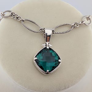 May include: A silver necklace featuring a square, emerald-green pendant. The pendant is set in a silver frame and hangs from a chain with oval links. The necklace is displayed on a neutral-colored surface.