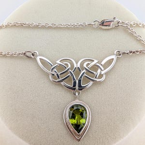 May include: A silver necklace featuring a polished metal chain and a central pendant with an intricate Celtic knot design. A single pear-shaped, faceted olive-green gemstone is suspended from the bottom of the knotwork, secured by a silver setting.