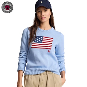 Op de afbeelding: Lichtblauwe trui met een Amerikaans vlagontwerp op de voorkant. De vlag heeft rode en witte strepen en een veld met witte sterren. De trui heeft lange mouwen en de letters "RL" zijn rood geborduurd.