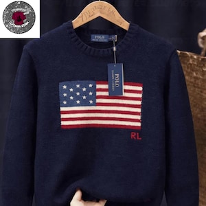 Puede incluir: Suéter azul marino con un diseño de la bandera estadounidense en la parte delantera. La bandera tiene estrellas blancas sobre un campo azul y rayas rojas y blancas. El suéter tiene cuello redondo y una etiqueta "POLO".