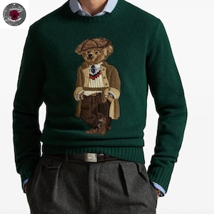 Puede incluir: Un suéter verde oscuro con un oso bordado que lleva sombrero y abrigo. El oso está representado en tonos marrones y beige. El suéter tiene cuello redondo y puños acanalados. El modelo lleva pantalones grises y un cinturón.