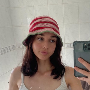 May include: A crochet bucket hat featuring horizontal stripes in shades of red, light pink, and a sage green trim along the edge. The hat is constructed with a textured yarn pattern and has a rounded crown with a downward sloping brim.
