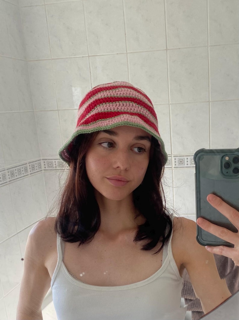 May include: A crochet bucket hat featuring horizontal stripes in shades of red, light pink, and a sage green trim along the edge. The hat is constructed with a textured yarn pattern and has a rounded crown with a downward sloping brim.
