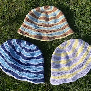 May include: Three handmade crochet bucket hats. The top hat features horizontal stripes in brown, light blue, and cream. The bottom left hat has dark blue and light blue stripes. The bottom right hat has alternating stripes of pale yellow and lavender.