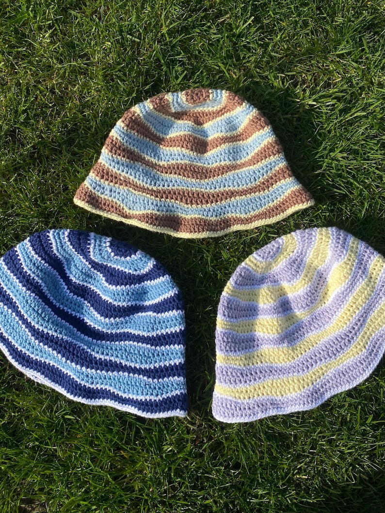 May include: Three handmade crochet bucket hats. The top hat features horizontal stripes in brown, light blue, and cream. The bottom left hat has dark blue and light blue stripes. The bottom right hat has alternating stripes of pale yellow and lavender.