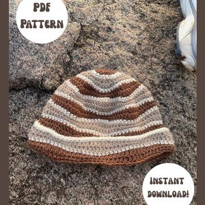 May include: A crochet bucket hat featuring horizontal stripes in shades of brown, beige, and cream. The image includes white circular text overlays that read PDF PATTERN and INSTANT DOWNLOAD! The hat is displayed on a textured stone surface.