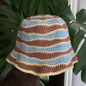 Puede incluir: Un sombrero de pescador tejido a ganchillo con rayas horizontales en marrón, azul claro y amarillo. El sombrero tiene una corona redondeada y un ala ligeramente acampanada. El sombrero es hecho a mano y parece estar hecho de hilo suave.