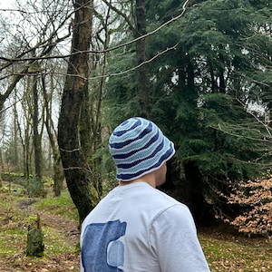 May include: A crochet bucket hat featuring concentric wavy stripes in shades of dark blue, medium blue, and light blue. The hat is worn with a white short-sleeved t-shirt that displays a large, abstract blue graphic print on the back.