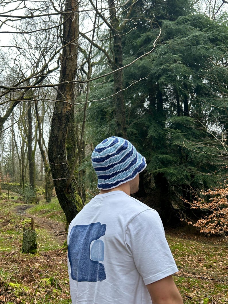 May include: A crochet bucket hat featuring concentric wavy stripes in shades of dark blue, medium blue, and light blue. The hat is worn with a white short-sleeved t-shirt that displays a large, abstract blue graphic print on the back.